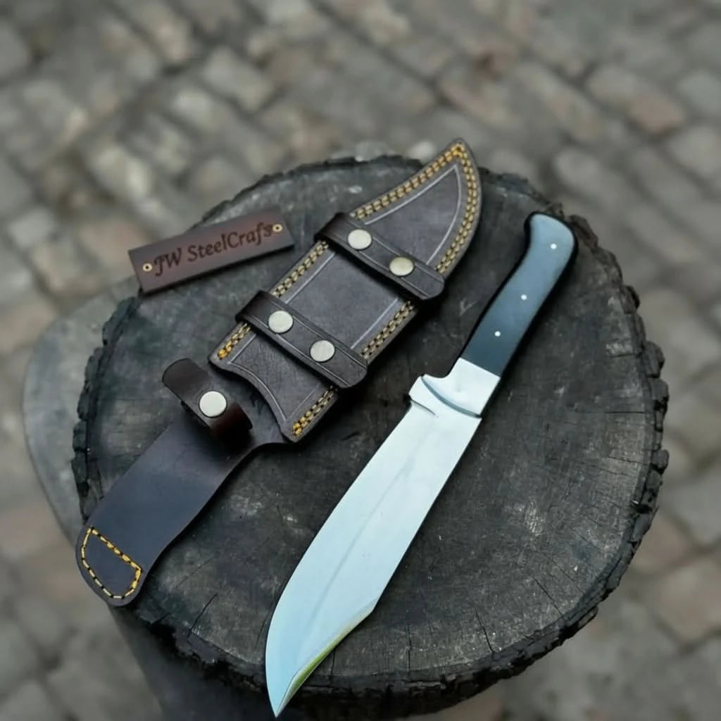 Rugged handmade fixed blade knife with black handle and custom leather sheath displayed on a wooden stump outdoors.