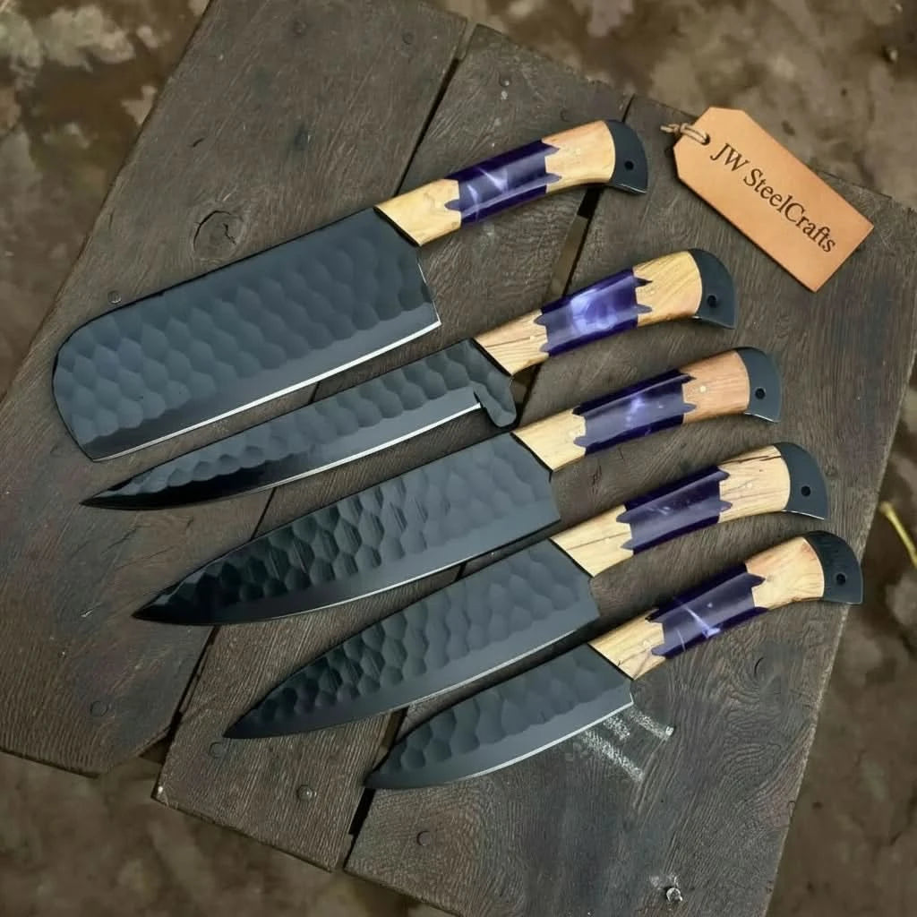 Angled overhead shot of handcrafted black hammered steel chef knives with purple resin and olive wood handles displayed in graduated sizes on a dark rustic wooden chopping board
