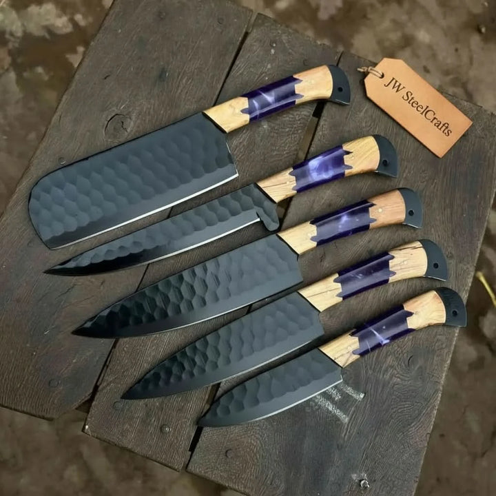 Angled overhead shot of handcrafted black hammered steel chef knives with purple resin and olive wood handles displayed in graduated sizes on a dark rustic wooden chopping board
