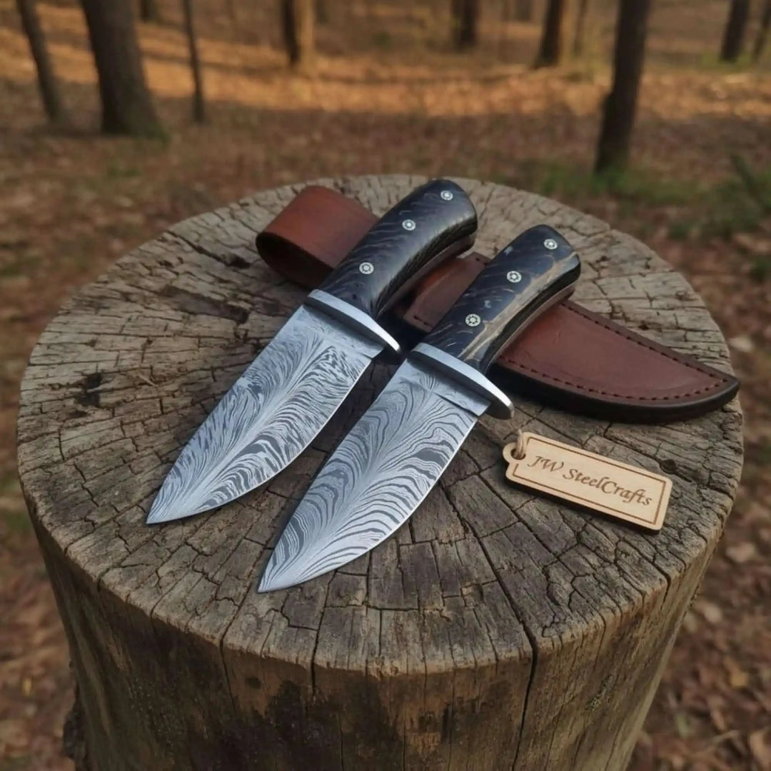Handmade Damascus knife set featuring two patterned blades, black handle scales, silver bolsters, and brown leather sheath outdoors.