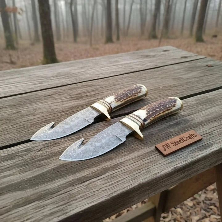 Polished Damascus hook blades in crossed display with stag handles, brass guards, and natural forest background.