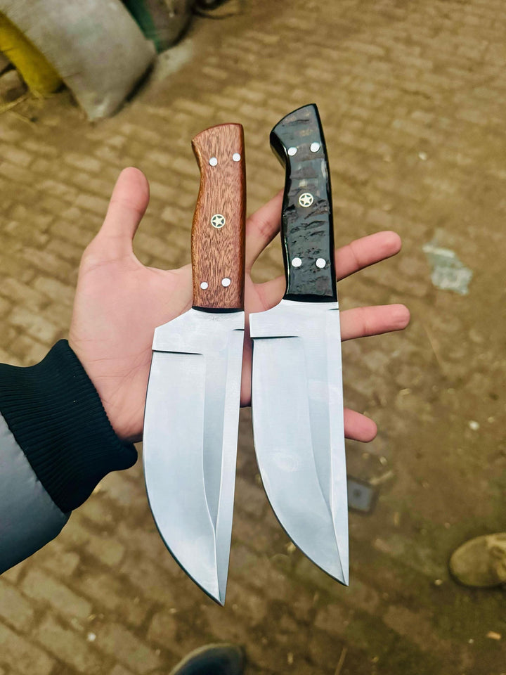 Two custom bowie-style knives with polished steel blades, one wood handle and one black handle, held in a hand outdoors.