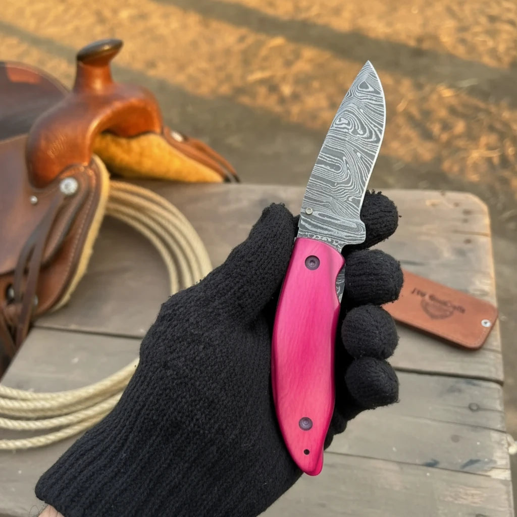 Pakka Wood Handmade Damascus Steel Folding Knife With Pink Handle Held By Gloved Hand On Wooden Surface