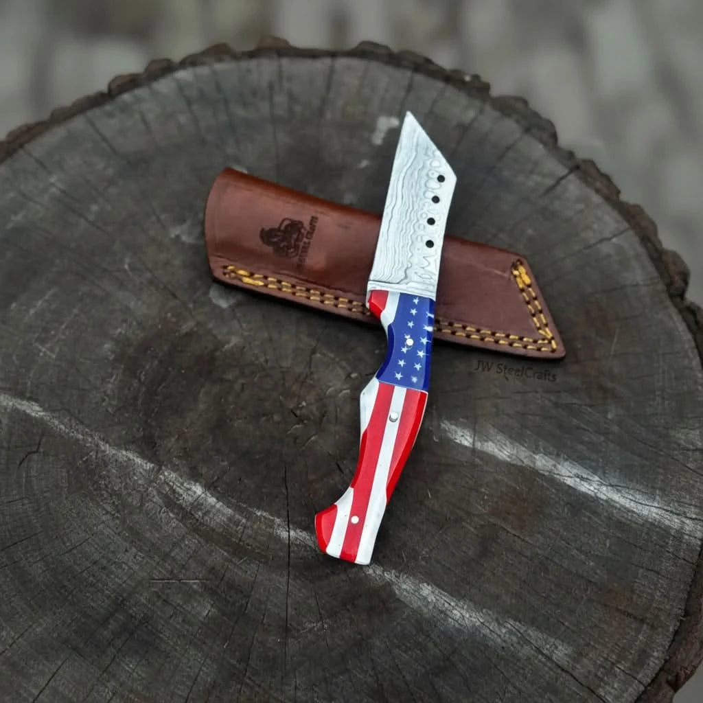 Red white blue handle Damascus knife with star design, layered steel blade, and brown leather sheath on rustic wood.