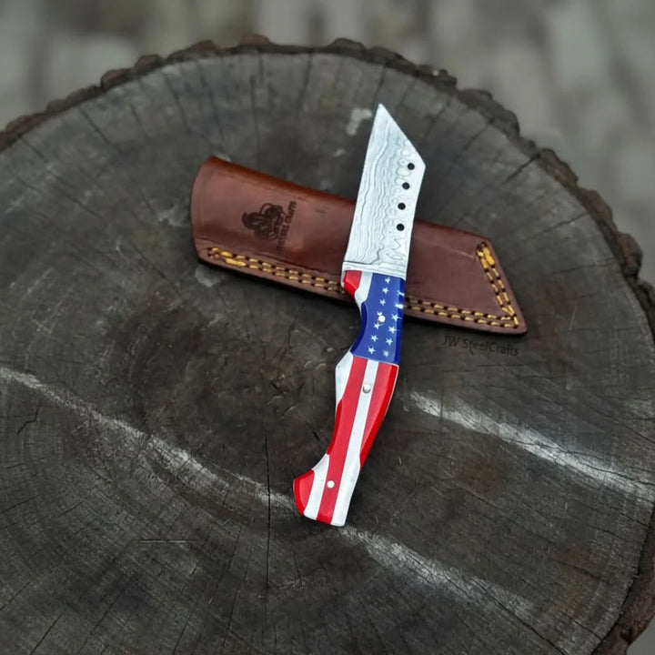 Red white blue handle Damascus knife with star design, layered steel blade, and brown leather sheath on rustic wood.