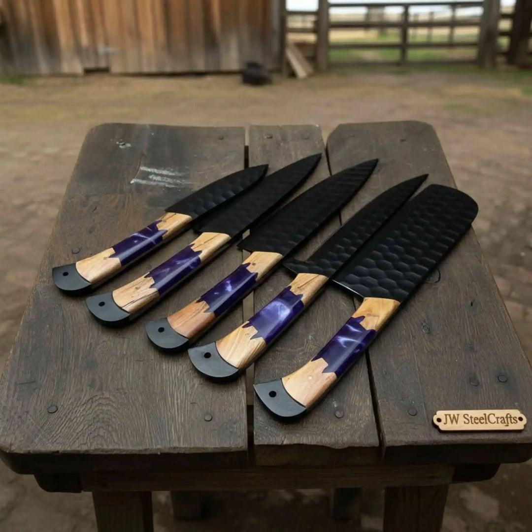 Five handcrafted black hammered finish kitchen knives with vivid purple epoxy resin and warm natural olive wood hybrid handles fanned out on a weathered wooden bench outdoors

