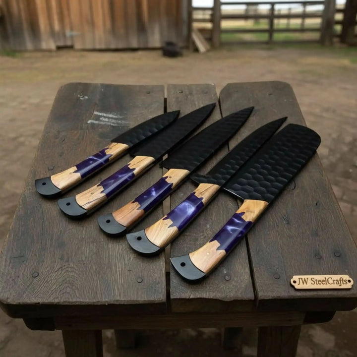 Five handcrafted black hammered finish kitchen knives with vivid purple epoxy resin and warm natural olive wood hybrid handles fanned out on a weathered wooden bench outdoors
