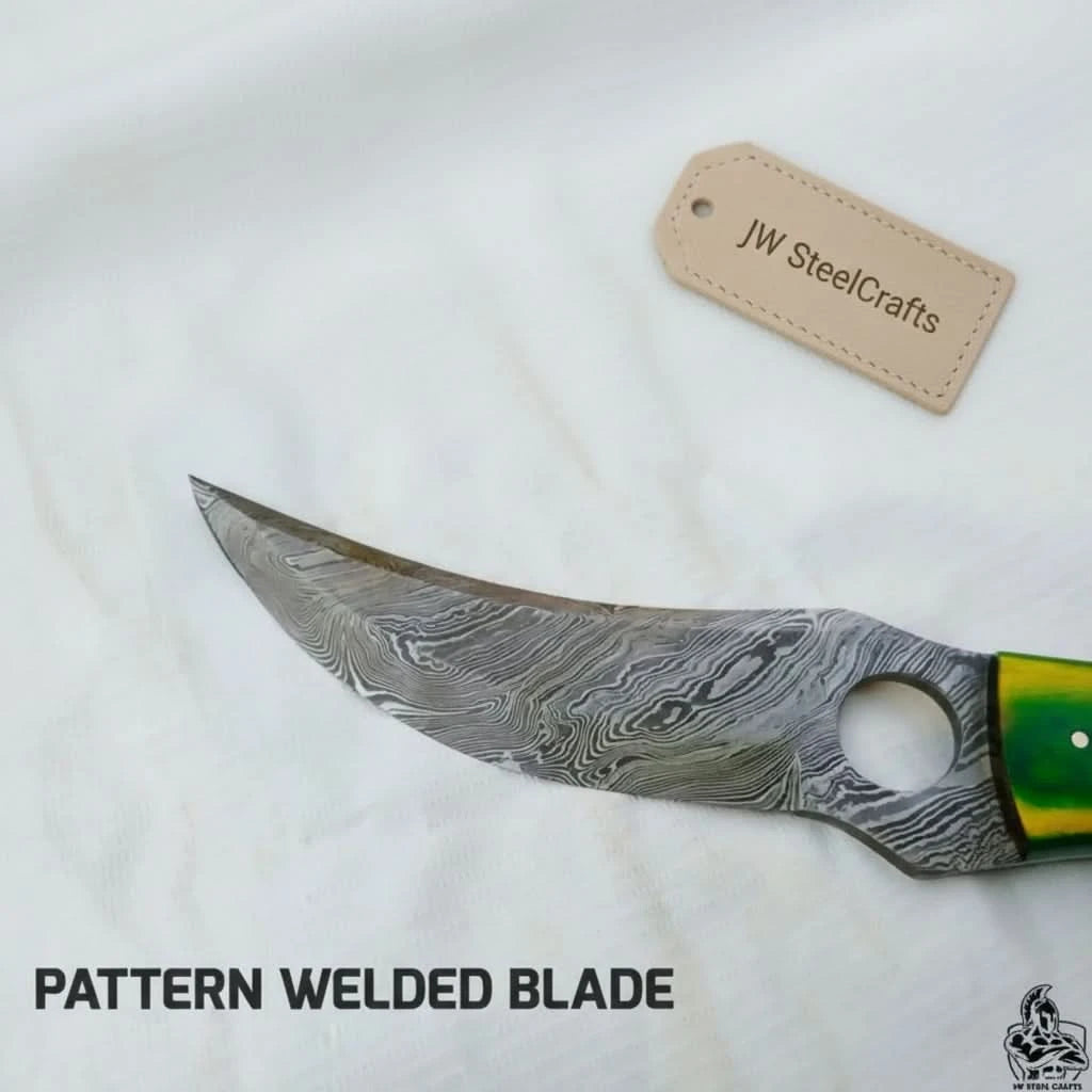 Sharp curved Damascus knife with finger hole, green yellow handle accent, cream grip, and forest stump background.