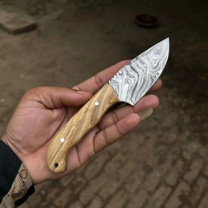 Premium Fixed Blade Damascus Steel Tracker Knife Held By A Hand, Featuring A Wooden Handle And Metallic Blade Against A Blurred Background.
