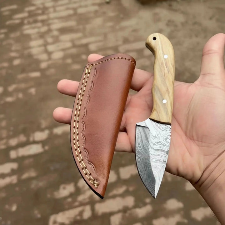 Premium Fixed Blade Damascus Steel Tracker Knife Held By A Hand, Featuring A Wooden Handle And Leather Sheath Against A Blurred Natural Background.