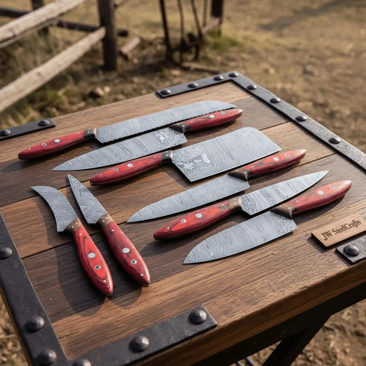Eight handcrafted Damascus steel kitchen knives with red wood handles displayed on rustic weathered wooden table outdoors.