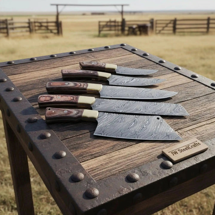 Five handcrafted Damascus steel kitchen knives with dark wood handles and brass bolsters displayed on rustic outdoor wooden table.