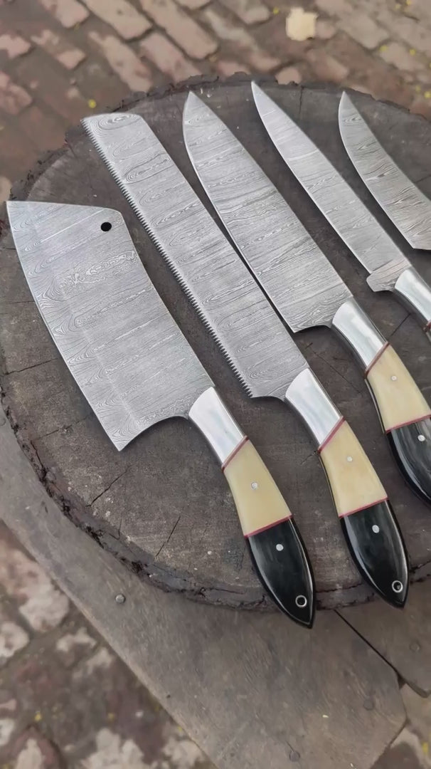 A cinematic showcase of a 5-piece handmade Damascus steel chef knife set. The video highlights the unique wavy blade patterns, premium cream and black resin handles, and the sharp edges of the cleaver and chef knives, all displayed in a rustic outdoor setting.