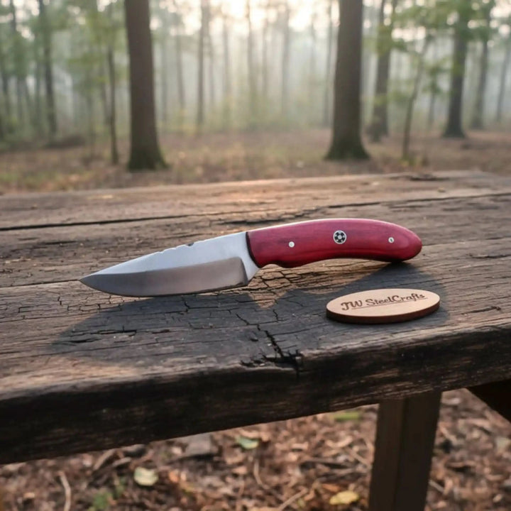 Handmade fixed blade knife featuring a red wood handle, polished drop point blade, white pins, and woodland background.