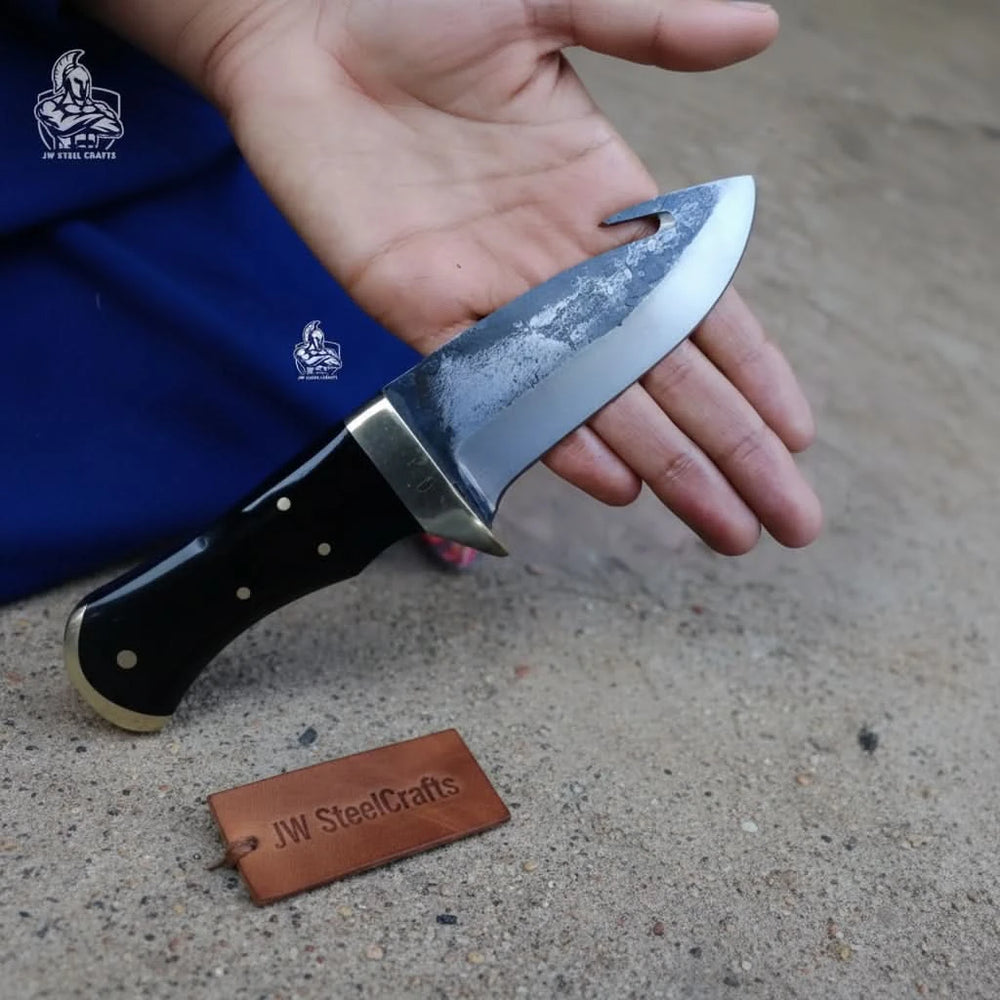 Bushcraft gut hook knife lying on a wooden table with forest trees and camping gear blurred in the background.