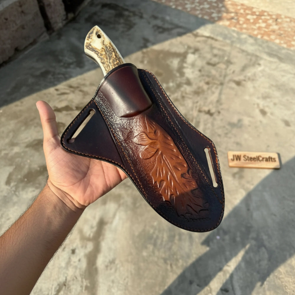 Embossed leather sheath holding a custom fixed blade knife with a stag-pattern handle in a person’s hand.