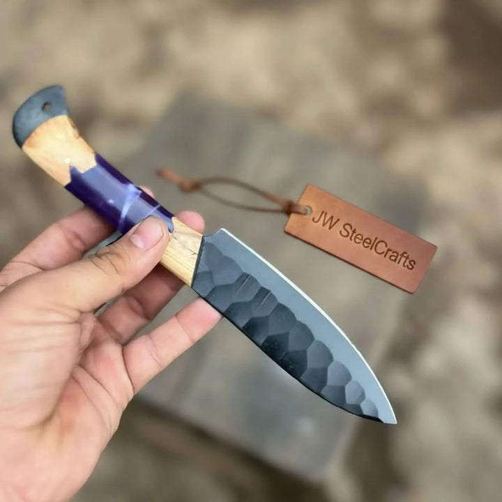 Detail shot of a handcrafted black hammered steel kitchen knife with vivid purple epoxy resin and warm olive wood hybrid handle held in hand outdoors
