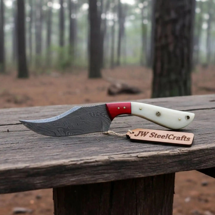 hunting knife with patterned steel blade, white handle, red bolster, and blurred pine forest background.