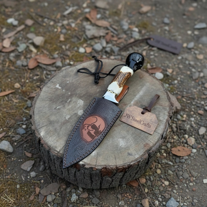 Stag handle fixed blade knife partially inserted into a stitched leather cover with a decorative skull panel.