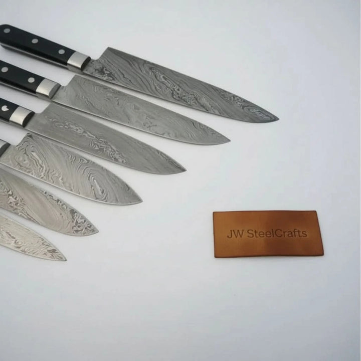 Closeup of six Damascus kitchen knife blades aligned diagonally against clean white studio background.