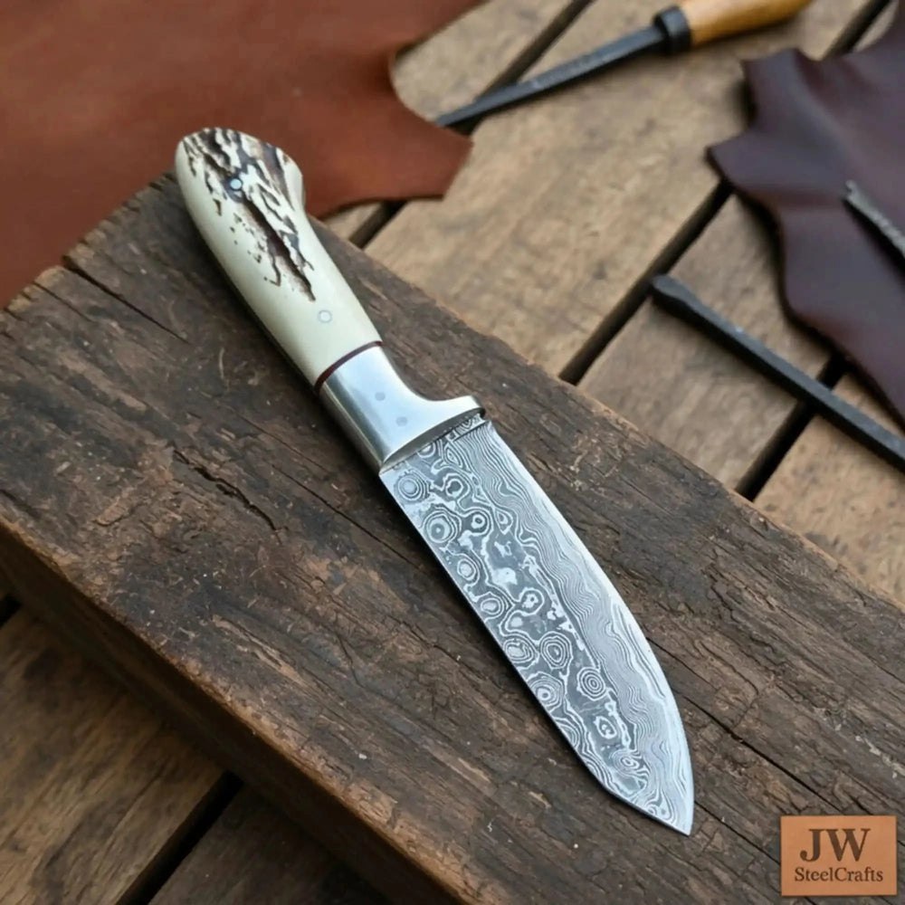 The Damascus Frostbite Skinner Knife Decorative Knife With A Detailed Blade Displayed On A Wooden Surface, Accompanied By Leather Tools.