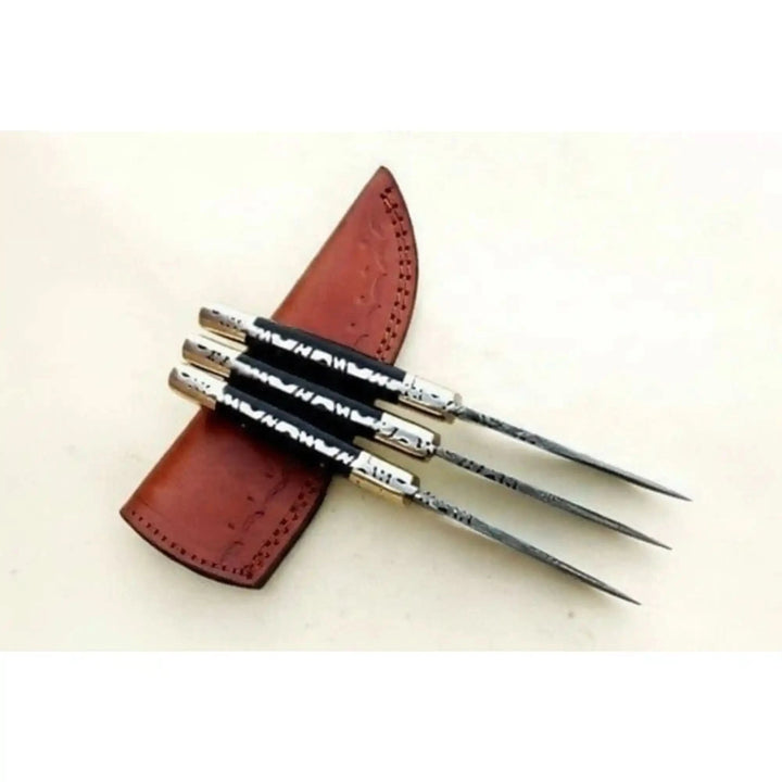 Handmade throwing knife set featuring three slim pointed blades, black grips, silver accents, and brown leather sheath