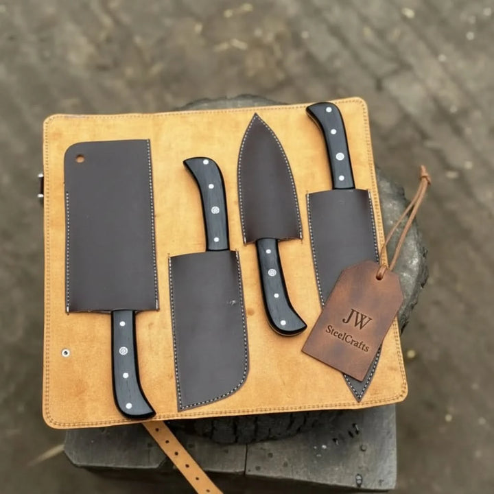 Four-piece chef knife set with black handles and individual dark brown stitched leather blade sheaths displayed on an open tan suede leather roll on a rustic anvil outdoors
