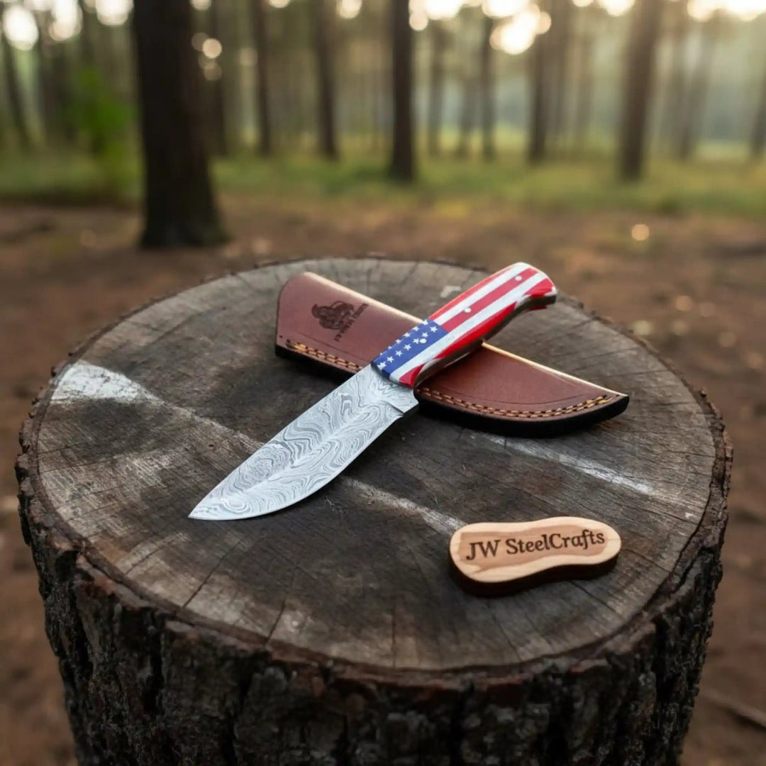Patriotic Damascus knife featuring red white and blue handle, patterned steel blade, brown leather sheath, and woodland background.