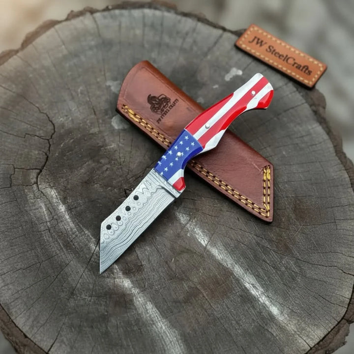 Damascus steel knife with USA flag handle, red white and blue grip, brown leather sheath, and tree stump display.