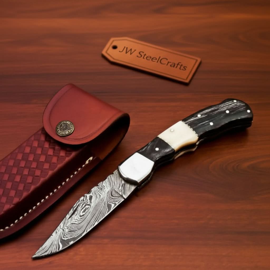     Ravenoak Damascus pocket knife with curved black grip and layered blade beside leather sheath