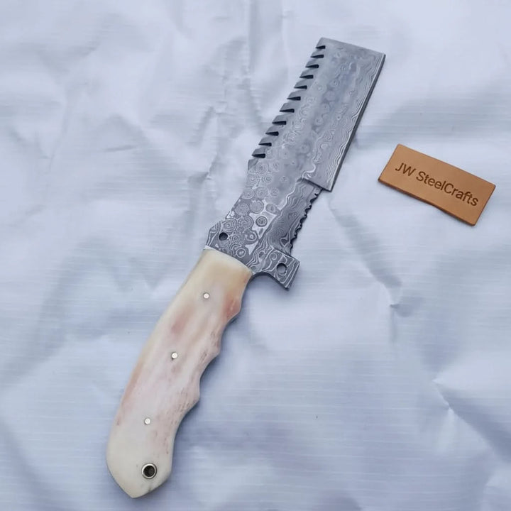 Serrated Damascus cleaver with an ivory handle displayed on a white cloth beside a JW SteelCrafts tag.