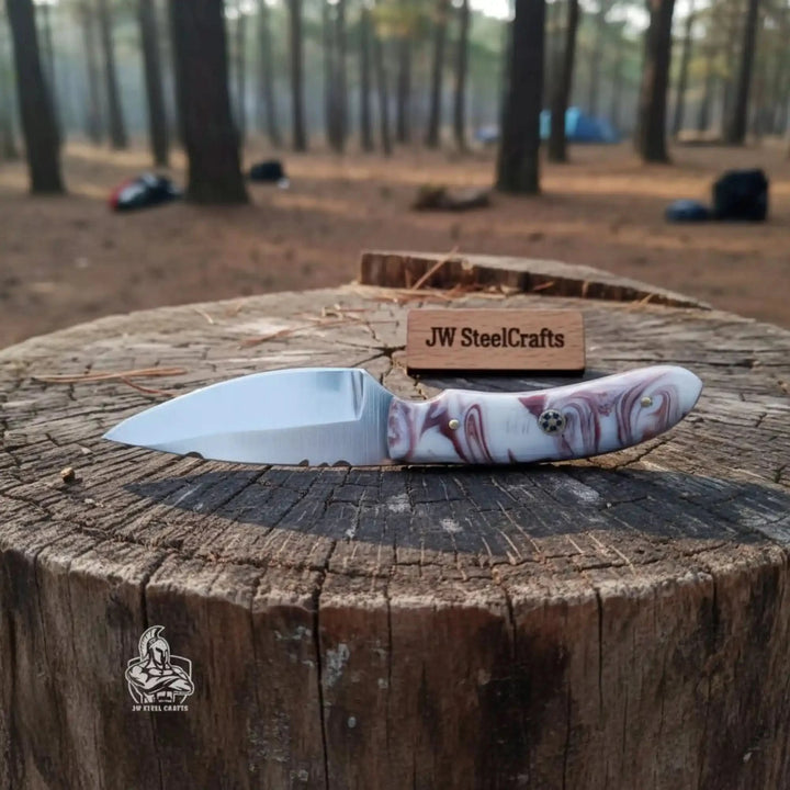 camping knife with marbled resin grip, polished blade, mosaic pin, and rustic forest stump background.