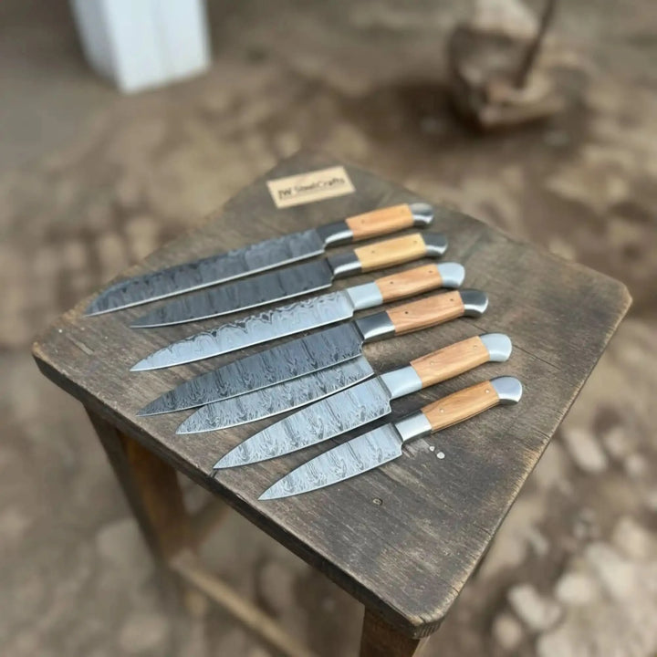 Eight-piece handmade Damascus steel kitchen knife set with olive wood handles displayed on a rustic wooden stool by JW Steel Craftes
