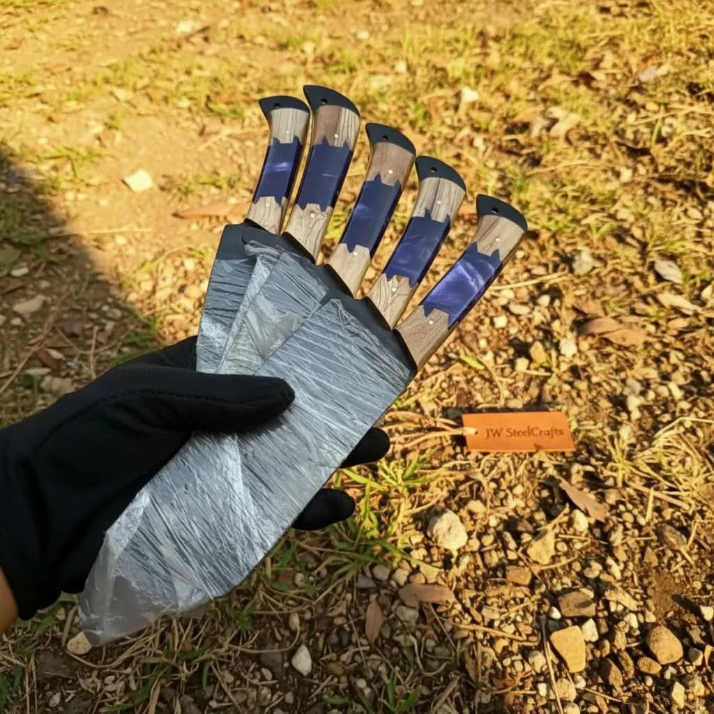 All five hand-forged black blade culinary knives with purple epoxy and olive wood handles fanned out and held together in a black gloved hand showing full set in one creative shot
